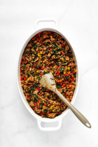 A white casserole dish filled with sausage crumbles, spinach, and yellow and red peppers. There is a spoon in the middle of it and the casserole dish is on a white counter.