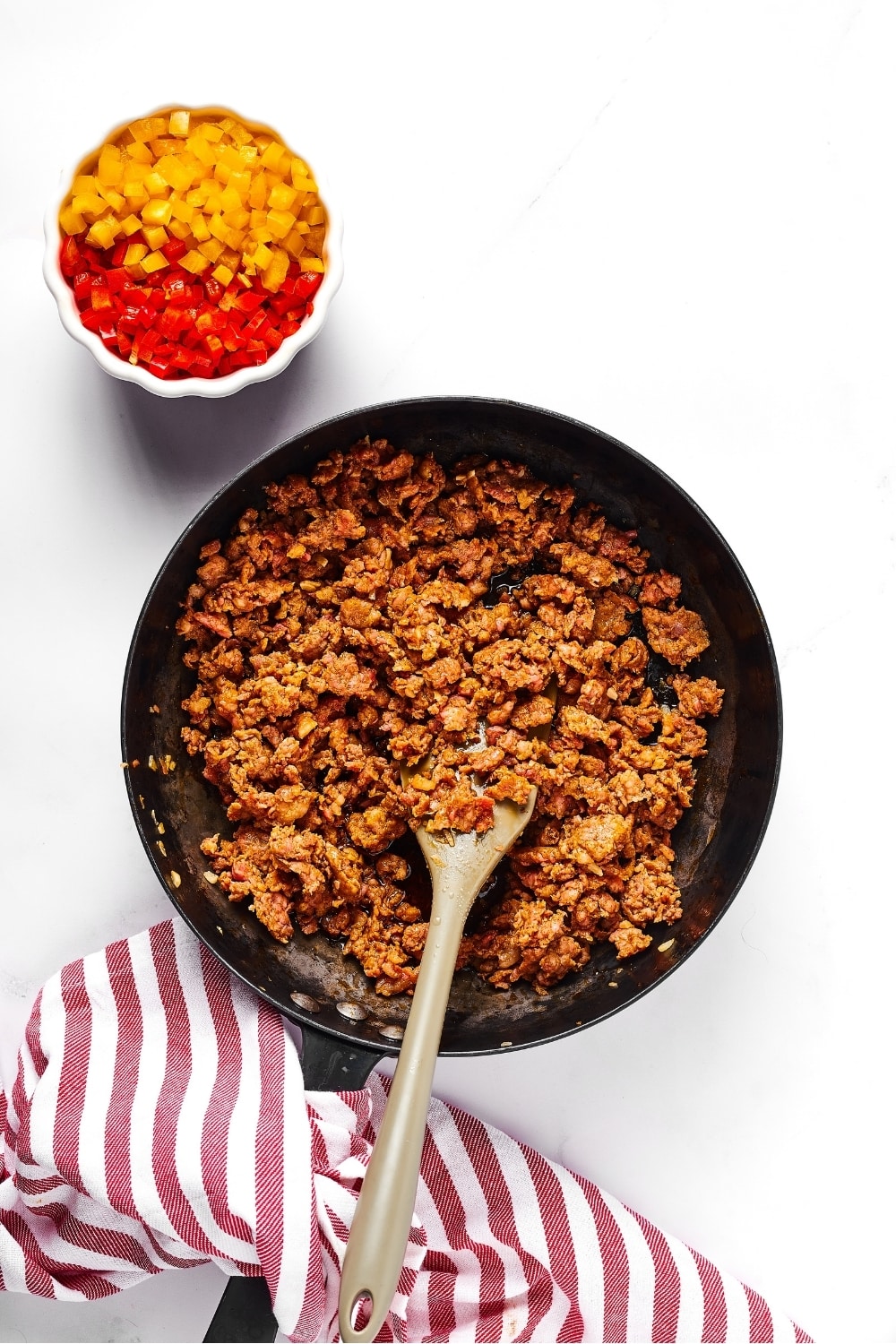 A saute pan with sausage crumbles in it. Behind the pan is a small white bowl with half red peppers and half yellow peppers.