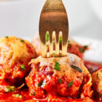 A gold fork puncturing a meatball. There is sauce in front of the meatball and a few meatballs around it.