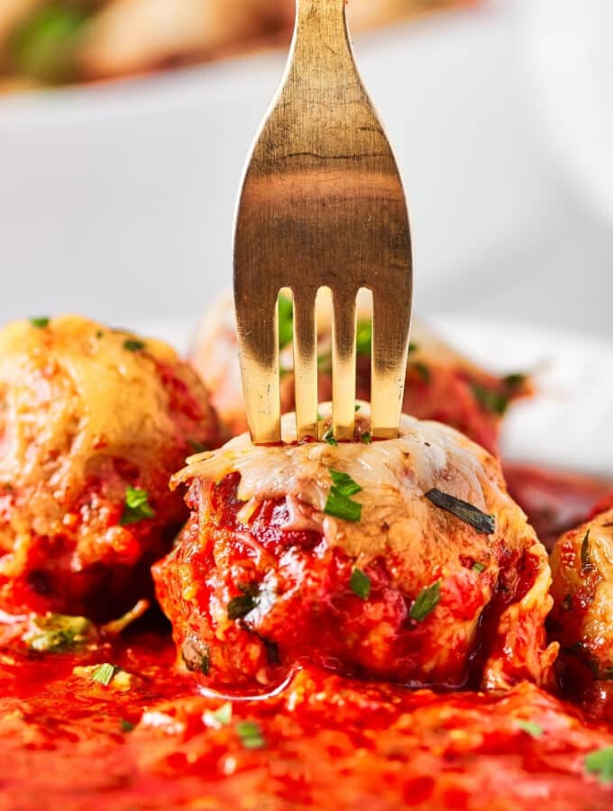 A gold fork puncturing a meatball. There is sauce in front of the meatball and a few meatballs around it.