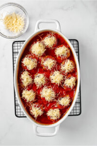 A white baking dish with meatballs in tomato sauce with shredded mozzarella cheese on top. The baking dish is on a wire rack on a white counter in behind it is a small glass bowl of mozzarella cheese.