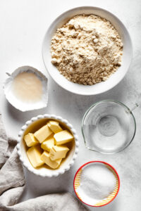 Ingredients for an almond flour pie crust. There is a small bowl of erythritol, a bowl of cubed butter, a small bowl of apple cider vinegar, a pitcher of water, and a large bowl of almond flour.