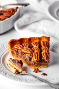 A slice of pecan pie and a white plate. A slice is taken out of the front of the pie and it is on a fork lane next to the left of the pie on the plate.