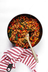 Black sauté pan with sausage crumbles, red and yellow peppers, and spinach with a wooden spoon in the middle of them. There is a red and white striped napkin on the handle.