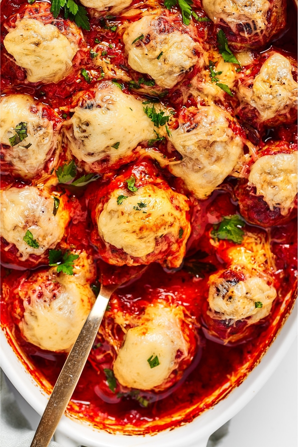 Part of the white baking dish filled with meatballs in tomato sauce with melted cheese on top.