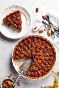 A pecan pie and a pie dish with a slice missing from it and a serving spatula where the slice was. Behind the pie is a white plate with a slice of pecan pie on it.