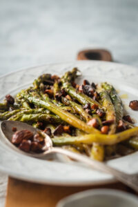 Asparagus on a white plate. There's a spoon with some almonds on it on the plate and some almonds on the asparagus.