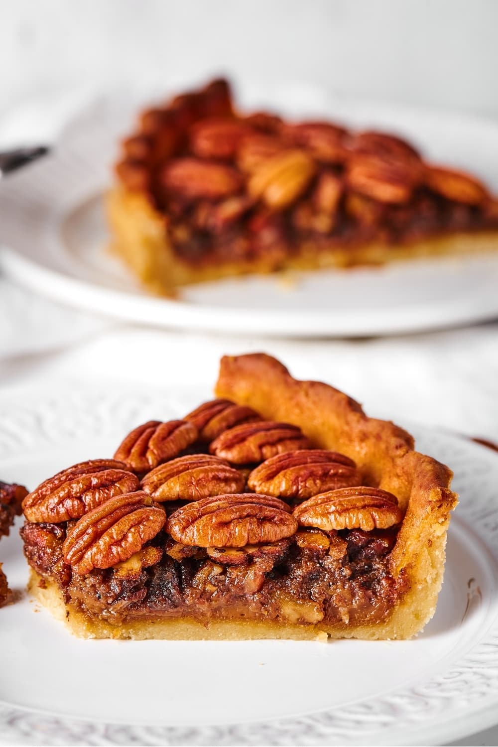 A Sideview of a slice of pecan pie on a white plate. Behind that is another slice of pecan pie on a white plate.
