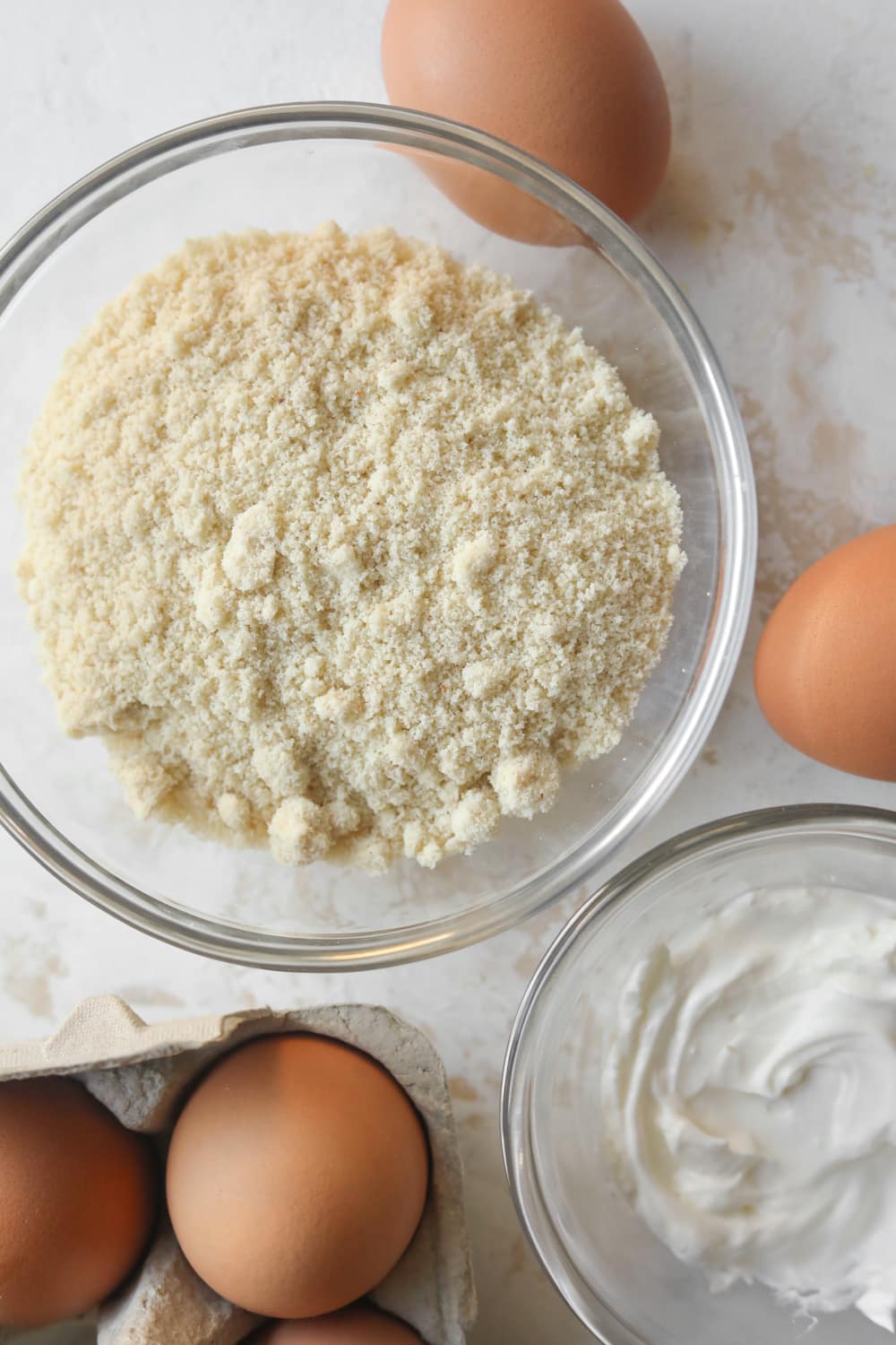 A glass bowl filled with almond flour, a glass bowl filled with sour cream, and brown eggs on a white table.