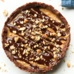 A peanut butter pie with a chocolate drizzle and crushed peanuts on top of it. The pie is set on a white table.