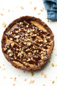 A peanut butter pie with a chocolate drizzle and crushed peanuts on top of it. The pie is set on a white table.