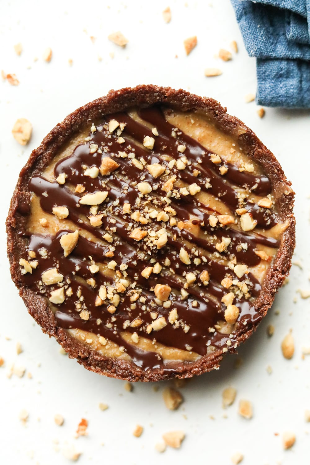 A peanut butter pie with a chocolate drizzle and crushed peanuts on top of it. The pie is set on a white table.