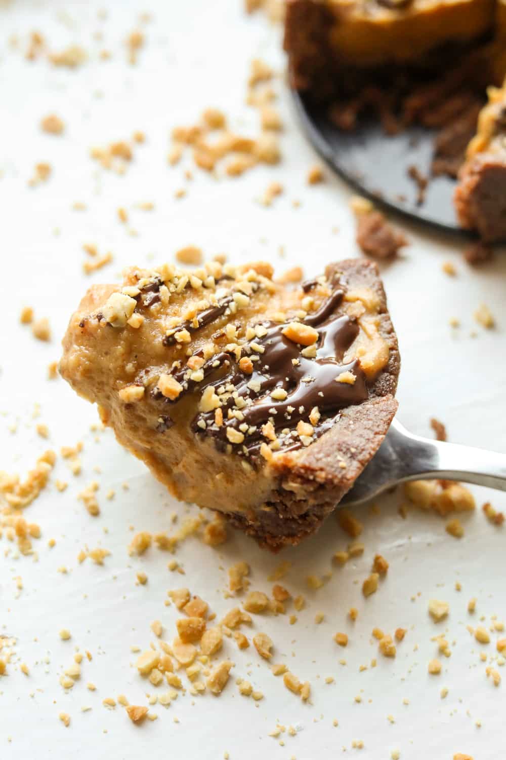 A piece of peanut butter pie on a fork set on a white table. There are crushed peanuts next to the piece of pie.