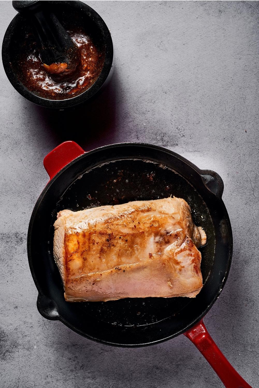 A seared pork loin in a pan with a red handle. The pan is on a white table.