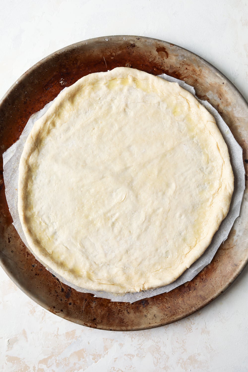 And uncooked pizza crust that's been brushed with olive oil set on a white sheet of parchment paper. The parchment paper is on top of a pizza tray.