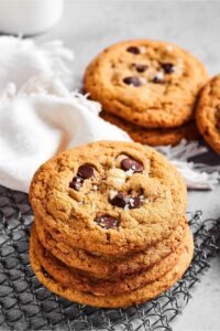 A stack of four chocolate chip cookies on a wire rack. Behind them is a white cloth with a few chocolate chip cookies behind it.