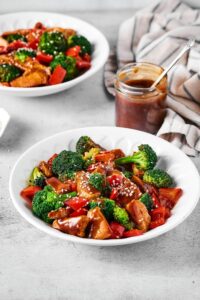 A white bowl filled with chicken stir fry with a glass cup of stirfry sauce behind it. Behind that is part of a white bowl filled with the stir fry.