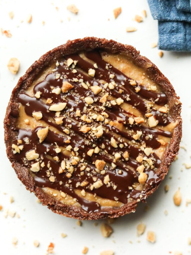 A peanut butter pie with a chocolate drizzle and crushed peanuts on top of it. The pie is set on a white table.