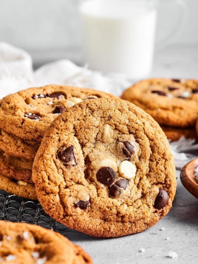 A chocolate chip cookie leaned up against a stack of chocolate chip cookies. Behind them is part of another cookie and a glass of milk.