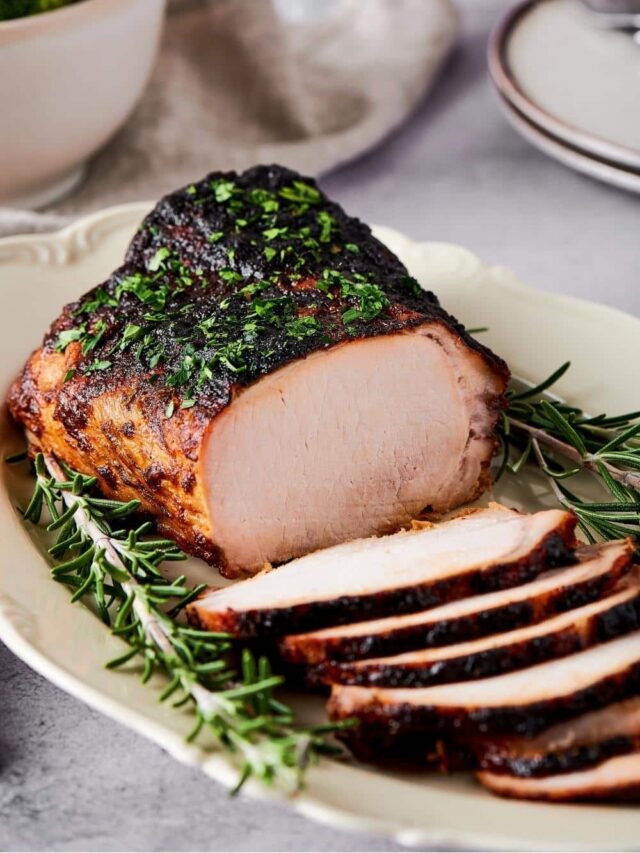 A white plate placed diagonally with a pork loin on it and a bunch of slices of the pork loin overlapping one another in front of it.
