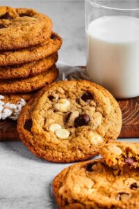 A chocolate chip cookie leaned up against a wooden cutting board with part of a stack of cookies on it and part of a glass of milk on it.