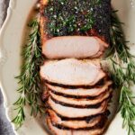 A whole pork loin on a white plate. There are six slices of pork loin's that are overlapping one another in front of the entire pork loin.