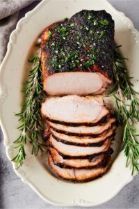 A whole pork loin on a white plate. There are six slices of pork loin's that are overlapping one another in front of the entire pork loin.