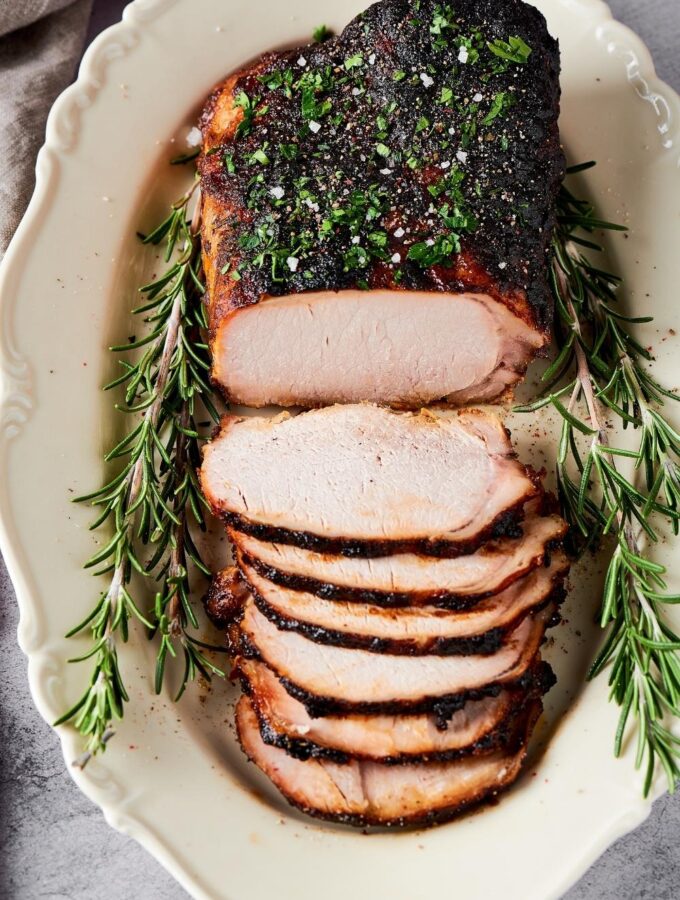 A whole pork loin on a white plate. There are six slices of pork loin's that are overlapping one another in front of the entire pork loin.