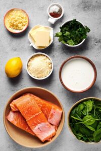 A bowl with three salmon fillets, a bowl of spinach, a bowl of heavy cream, a small bowl of Parmesan cheese, lemon, edition butter, small bowl of parsley, small bowl of minced garlic, and a small bowl of seasonings all on a gray counter.