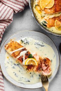 A salmon fillet on top of a garlic butter sauce covering a gray plate. Behind it is a quarter of a skillet with the salmon and sauce in it.