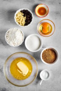 A glass bowl of melted butter, a cup of brown sugar, a cup of sugar, a bowl of flour, a bowl with two eggs, a bowl with half white chocolate chips half milk chocolate chips, and a bowl of vanilla extract all on a gray counter.