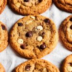 Three chocolate chip cookies in a row on a white tablecloth with parts of chocolate chip cookies on both sides of them.