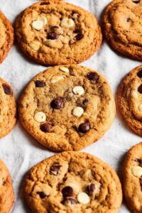 Three chocolate chip cookies in a row on a white tablecloth with parts of chocolate chip cookies on both sides of them.