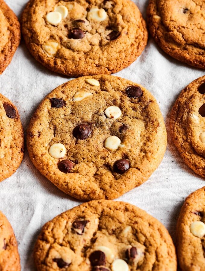 Three chocolate chip cookies in a row on a white tablecloth with parts of chocolate chip cookies on both sides of them.