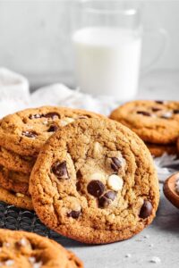 A chocolate chip cookie leaned up against a stack of chocolate chip cookies. Behind them is part of another cookie and a glass of milk.
