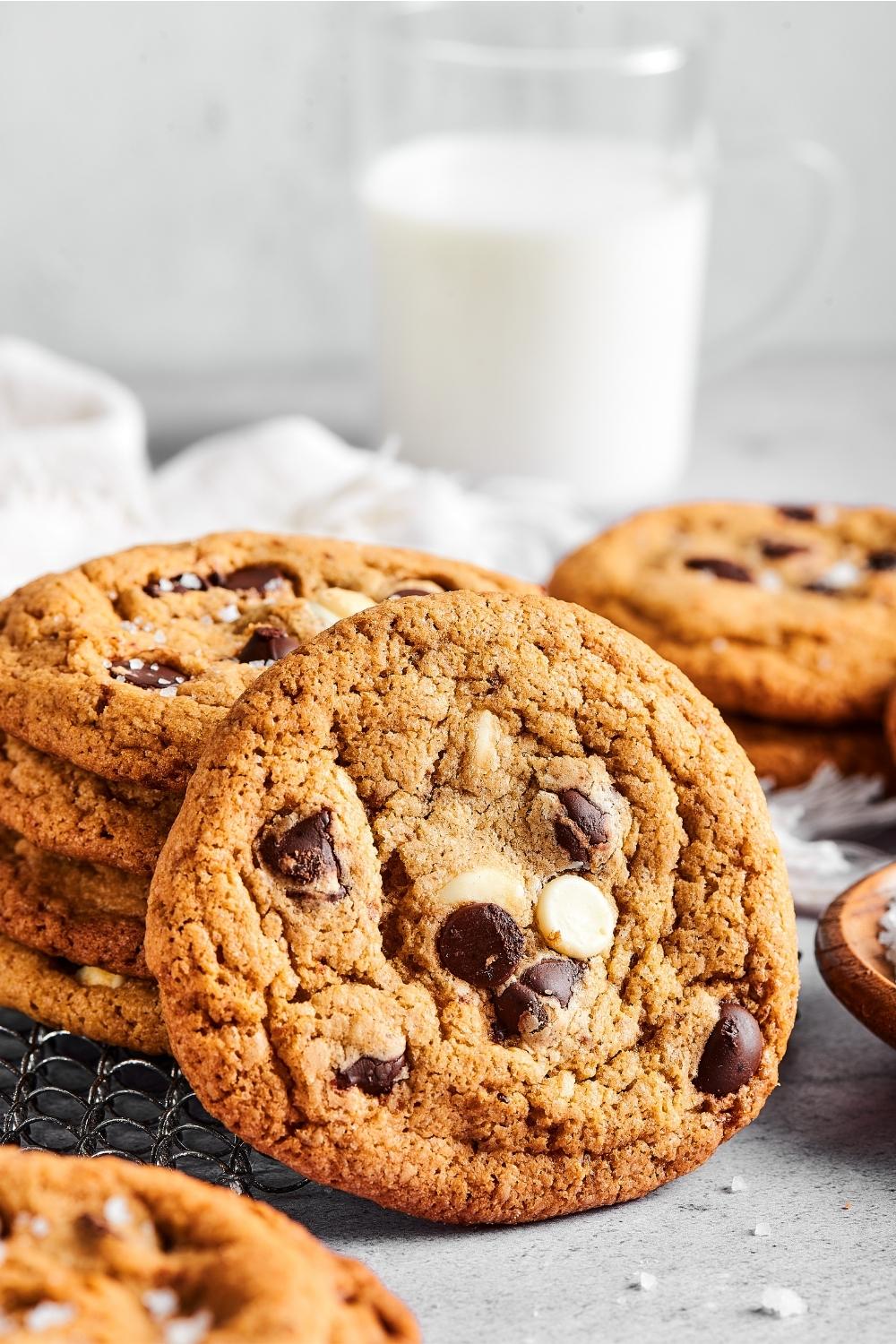 A chocolate chip cookie leaned up against a stack of chocolate chip cookies. Behind them is part of another cookie and a glass of milk.