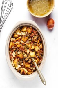 A casserole dish filled with stuffing. Behind it is part of a bowl of chicken broth with a cracked egg shell in front of it.