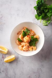 Shrimp in a white bowl that is on a gray counter. There's part of a bowl cilantro in front of it and two lemon wedges to the side of it.