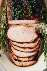 Heart of a pork loin roast with six slices overlapping one another that are cut out from the part of the pork loin. The pork loin is on a white plate and surrounded by sprigs of Rosemary.