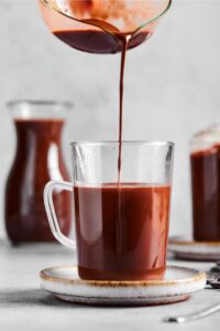 Part of a picture pouring hot chocolate into a glass cup that is ¾ of the way filled with hot chocolate.