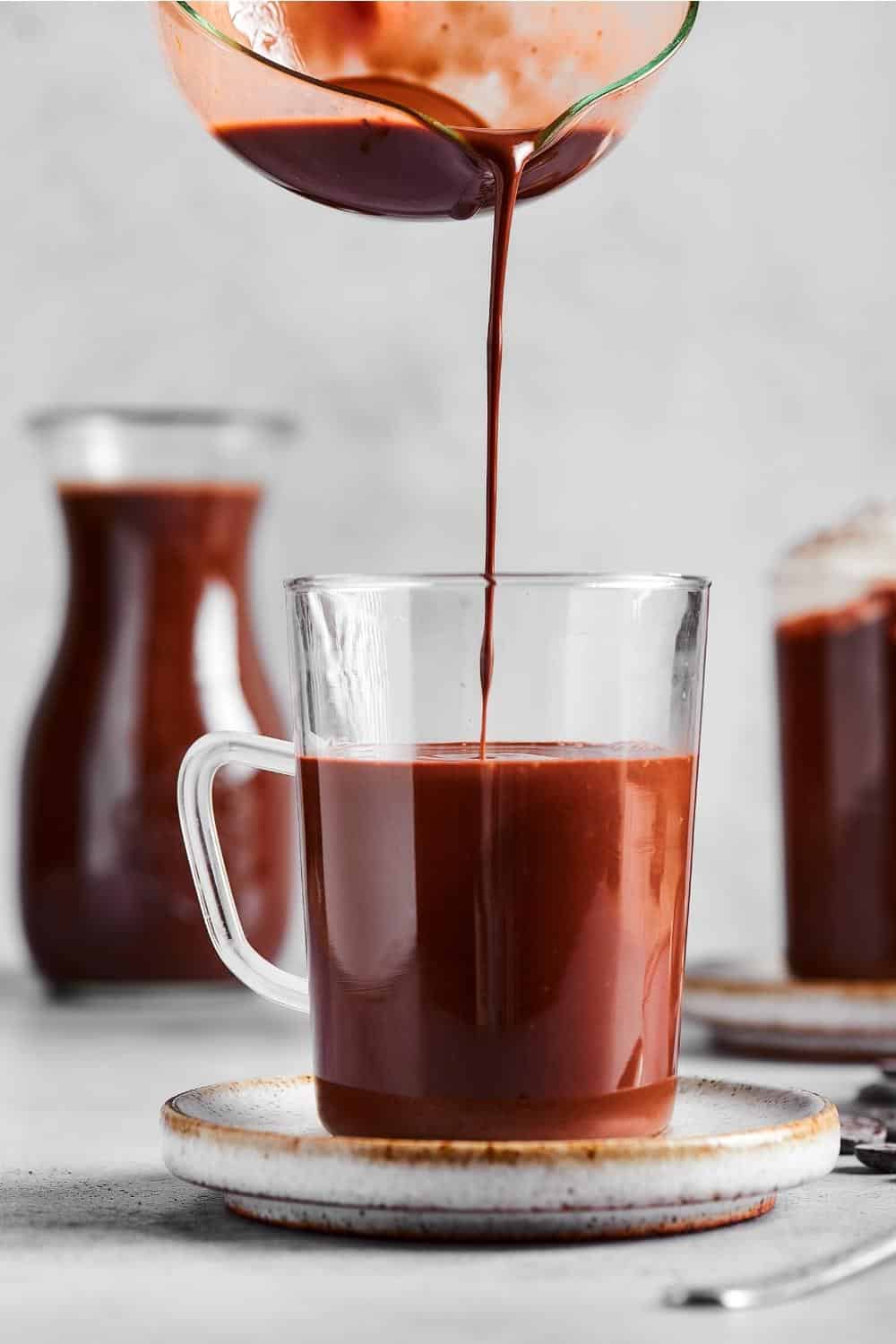 Part of a picture pouring hot chocolate into a glass cup that is ¾ of the way filled with hot chocolate.