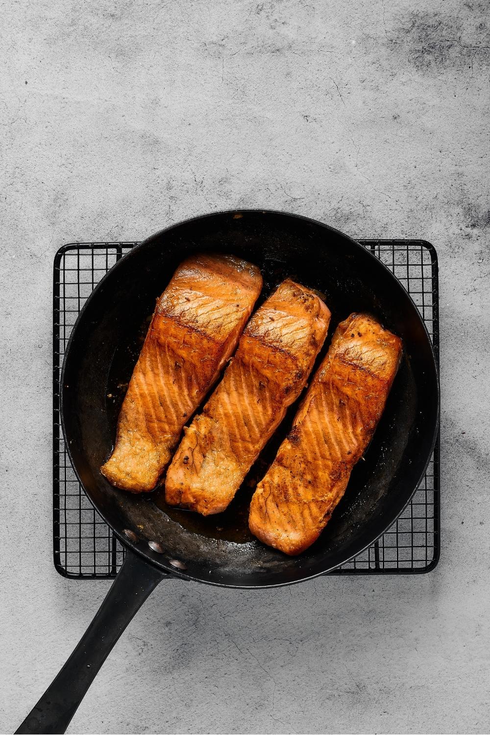 Black skillet filled with three salmon fillets. The skillet is on a wire rack on a gray counter.