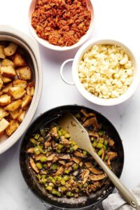 A skillet with chopped mushrooms and peppers, a white bowl filled with cauliflower, part of a white bowl with sausage crumbles, and part of a casserole dish with toasted bread slices in it all on a white counter.