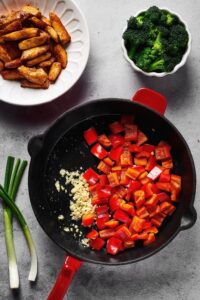 Skillet filled with sliced red bell peppers and minced garlic. Behind the skillet is a cup of broccoli and part of a plate with cooked chicken on it.