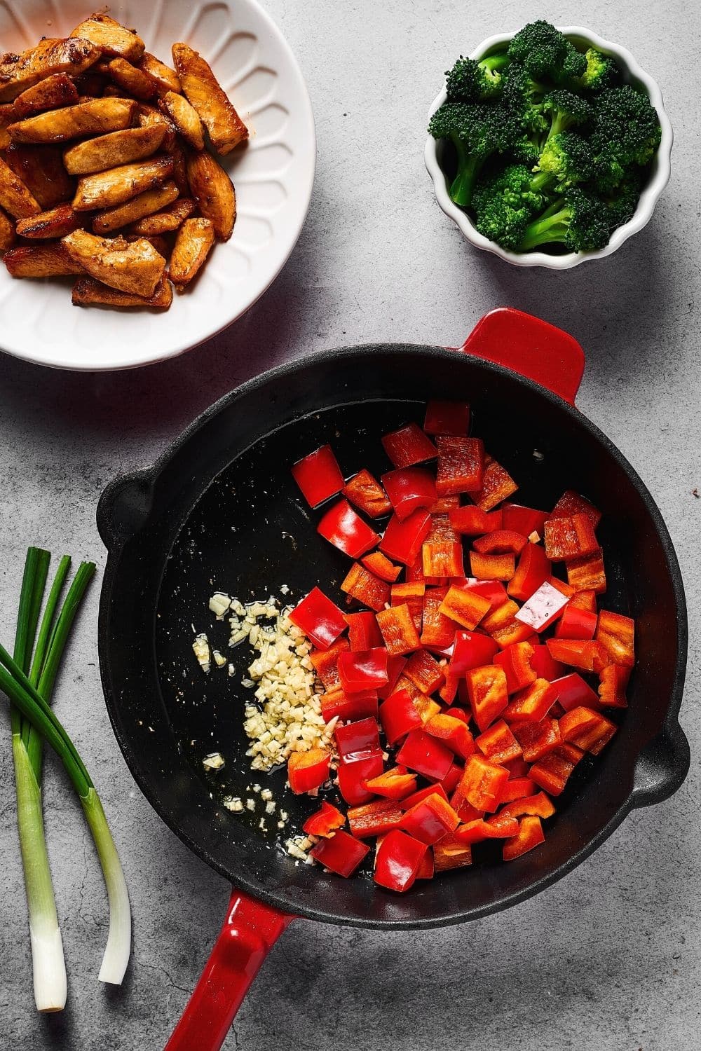 Skillet filled with sliced red bell peppers and minced garlic. Behind the skillet is a cup of broccoli and part of a plate with cooked chicken on it.