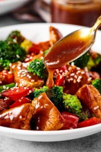 Part of a white bowl filled with chicken stir fry. There is a spoon pouring stir fry sauce onto the stir fry.