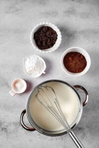 A pot filled with milk with a whisk in it, a small bowl of salt, a small bowl of confectioner erythritol, a small bowl of dark chocolate pieces, and a small bowl of cocoa powder on a gray counter.