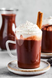 A glass mug filled with hot chocolate with whip cream on top and two cinnamon sticks in it. The mug is on a small gray plate in behind it is a glass pitcher of hot chocolate.