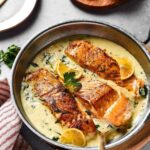 A skillet on a wood board filled with garlic butter sauce and three salmon fillets. Behind it is part of a wood cutting board with lemon wedges on it.