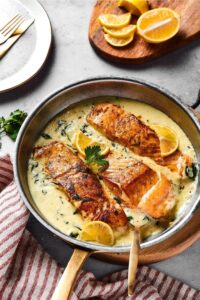 A skillet on a wood board filled with garlic butter sauce and three salmon fillets. Behind it is part of a wood cutting board with lemon wedges on it.